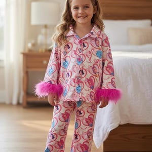 May include: A young girl wearing a pink pajama set with a princess-themed pattern. The pajamas have long sleeves and pants, trimmed with bright pink feather cuffs. A matching headband completes the outfit. The background includes a bedroom setting.