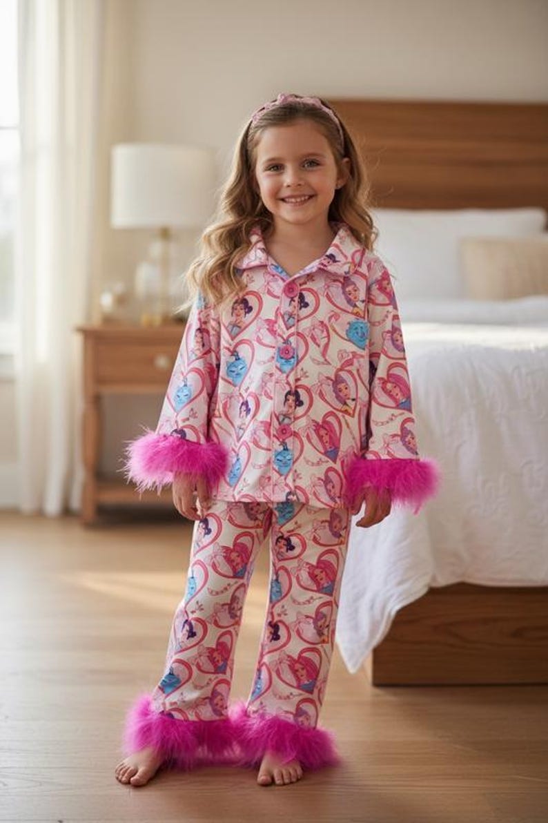 May include: A young girl wearing a pink pajama set with a princess-themed pattern. The pajamas have long sleeves and pants, trimmed with bright pink feather cuffs. A matching headband completes the outfit. The background includes a bedroom setting.