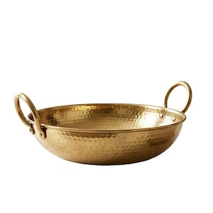 May include: A round, shallow, hammered brass bowl with two curved handles on opposite sides. The exterior and interior surfaces feature a distinct dimpled texture, reflecting light across the metallic gold-toned surface of the vessel.