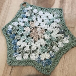 May include: A handmade, star-shaped crochet potholder in shades of green, white, blue, and tan. The potholder features a floral pattern in the center and a loop for hanging. The item is made of yarn.
