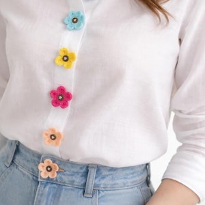 Op de afbeelding: Wit overhemd met gehaakte bloemknoppen in blauw, geel, roze en perzik. Het overhemd wordt gecombineerd met een lichtblauwe jeans. De kleurrijke bloemknoppen geven het klassieke overhemd een speelse uitstraling.