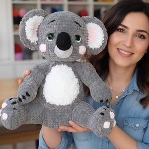 Puede incluir: Un peluche de koala gris hecho a mano con vientre blanco, detalles rosas en las orejas y mejillas, y ojos verdes. El koala tiene una nariz negra y está hecho de material suave y texturizado. Un lindo peluche.