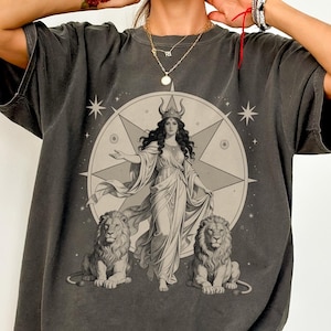 Könnte beinhalten: Dunkelgraues T-Shirt mit einer detaillierten Illustration einer Frau in einem fließenden Kleid, flankiert von zwei Löwen, vor einem Sternmuster. Das Wort "PEPPER" ist unten aufgedruckt. Das Shirt hat kurze Ärmel und eine lockere Passform.