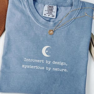 Puede incluir: Una camiseta azul vaquero con el texto "Introvert by design, mysterious by nature". Un collar dorado con un colgante está drapeado sobre la camiseta. La camiseta tiene cuello redondo y una etiqueta Comfort Colors.