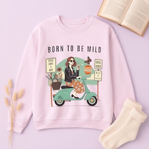 May include: Light pink sweatshirt with the text "BORN TO BE MILD". The graphic features a woman on a teal scooter, holding a drink, with signs indicating "Chill Vibes" and a speed limit of 5 MPH. A butterfly and flowers are also included.