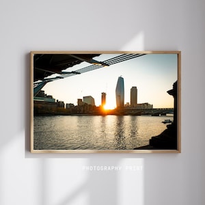 Può includere: Una fotografia di una città al tramonto, scattata da un ponte. Il sole tramonta dietro gli edifici, proiettando un bagliore dorato sulla scena. Gli edifici sono in silhouette contro il cielo, e il fiume in primo piano riflette la luce.