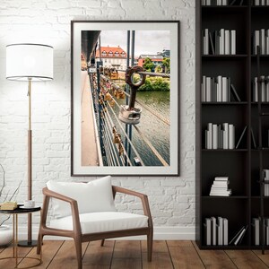May include: A framed photograph of a bridge with a river flowing underneath. The bridge is decorated with many padlocks, some of which are heart-shaped. The padlocks are attached to a metal railing.