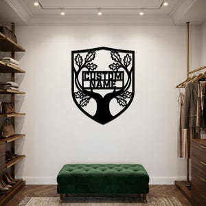 May include: A black metal wall sign shaped like a shield featuring a tree silhouette with oak leaves and a central rectangular cutout containing the text CUSTOM NAME. The design is symmetrical with branches extending upward and outward.