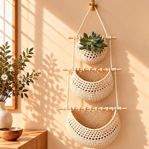 May include: A three-tiered wall hanging organizer featuring cream-colored crocheted baskets suspended by light-toned wooden dowels and thick cord. The top basket contains a green succulent plant, while the two lower baskets remain empty.