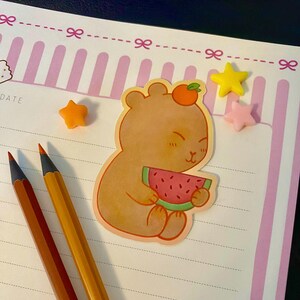 Peut inclure: Un autocollant mignon représentant un ours brun tenant une tranche de pastèque, surmonté d'une orange. L'autocollant est sur un bloc-notes à rayures roses et blanches, avec des ornements en forme d'étoiles orange, jaunes et roses et deux crayons.