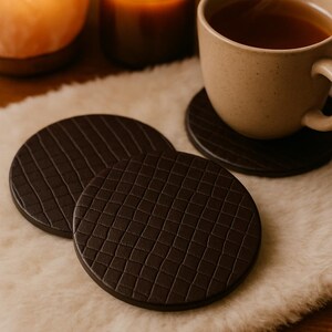 May include: A cozy scene featuring dark brown leather coasters with textured patterns. A beige mug of tea sits on one coaster, with a lit candle and a salt lamp in the background. The coasters are on a fluffy white surface.