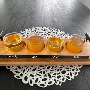 May include: A wooden beer flight with four glasses filled with golden beer. The tray has black handles and labels that read "Lager", "Ale", "Lager", and "Beer". The tray sits on a white lace doily.
