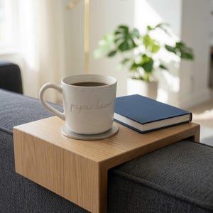 May include: A light-colored wooden sofa arm tray with a flat top and two downward-extending sides. Resting on the tray is a speckled white ceramic mug with the text "papa bear" in cursive script, a circular coaster, and a small navy blue book.