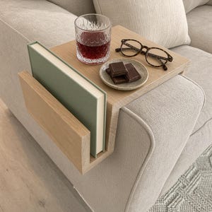 May include: A light-toned wooden sofa arm table featuring a flat top surface and a side pocket. The table holds a clear glass with a dark red liquid, a small speckled plate with three chocolate squares, a pair of tortoiseshell glasses, and a book.