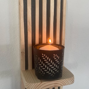 May include: A wall-mounted wooden sconce featuring vertical light-colored wood slats over a black backing. A small, black metal candle holder with a leaf-pattern cutout design sits on the shelf, containing a white candle with a lit flame.