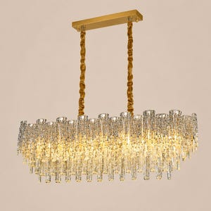 May include: A rectangular chandelier with a gold-colored frame and hanging rods. The light fixture is adorned with numerous clear, crystal-like elements, creating a sparkling effect. The overall design is elegant and suitable for interior lighting.