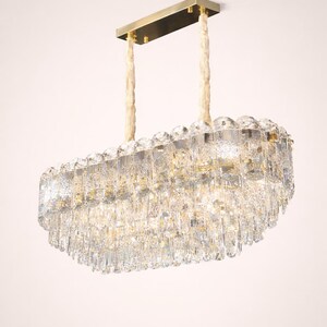 May include: A rectangular crystal chandelier with a gold-toned metal frame and hanging accents. The light fixture is suspended by two cream-colored rope-like cords. The crystals are arranged in layers, creating a cascading effect.