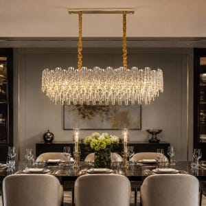 May include: A rectangular chandelier featuring multiple tiers of clear, vertical glass rods suspended from a gold-toned metal frame. The fixture is attached to the ceiling by two gold-toned chains and emits a warm, ambient light throughout.