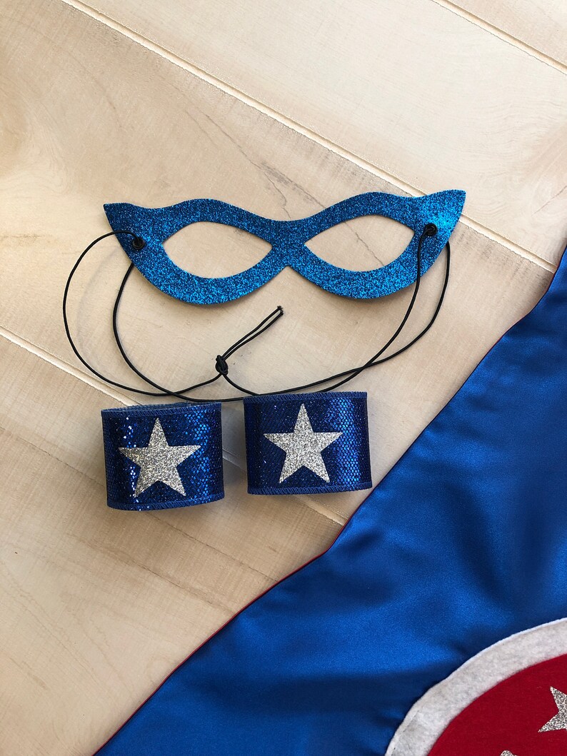 Puede incluir: Un conjunto de disfraz de superh&eacute;roe azul con una m&aacute;scara de ojos brillante, dos pu&ntilde;os azules con estrellas plateadas y una capa azul con un c&iacute;rculo rojo y blanco.