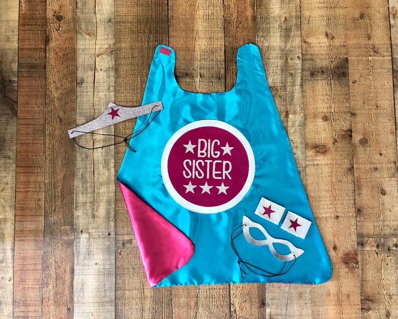 Puede incluir: Una capa de superh&eacute;roe azul y rosa con una estrella plateada y las palabras "Big Sister" en un c&iacute;rculo rosa. La capa viene con una corona plateada, una m&aacute;scara plateada y dos parches de estrellas plateadas.