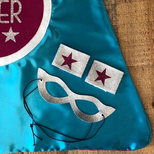 Puede incluir: Una capa de superh&eacute;roe turquesa con un c&iacute;rculo rojo y un borde blanco que dice "Super Star" con estrellas plateadas brillantes. La capa viene con una m&aacute;scara de ojos de brillo plateado y dos brazaletes de brillo plateado con estrellas rojas brillantes.