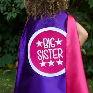 Puede incluir: Capa de superh&eacute;roe morada y rosa con un c&iacute;rculo blanco y el texto "BIG SISTER" en letras plateadas brillantes.