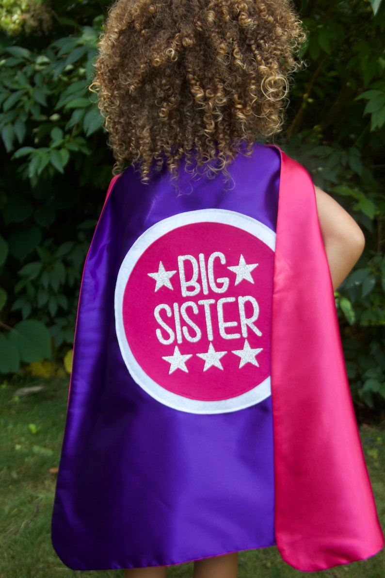 Puede incluir: Capa de superh&eacute;roe morada y rosa con un c&iacute;rculo blanco y el texto "BIG SISTER" en letras plateadas brillantes.