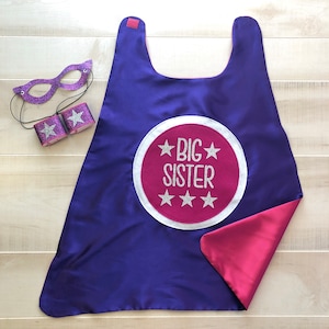 Puede incluir: Capa de superh&eacute;roe morada y rosa con un c&iacute;rculo rosa en la parte posterior que dice "BIG SISTER" en letras plateadas con purpurina y estrellas plateadas con purpurina alrededor del texto. La capa viene con una m&aacute;scara para los ojos morada y rosa a juego y dos brazaletes.