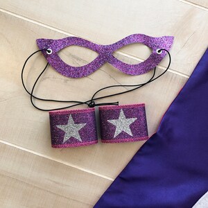 Puede incluir: Conjunto de disfraz de superh&eacute;roe de purpurina morado y rosa con m&aacute;scara, pu&ntilde;os y capa. La m&aacute;scara tiene una banda el&aacute;stica negra. Los pu&ntilde;os tienen estrellas de purpurina plateadas. La capa es morada.