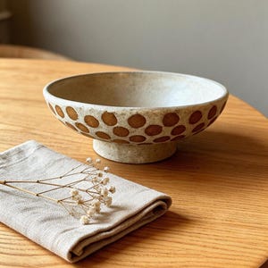 May include: A speckled off-white ceramic bowl with a pedestal base, featuring a repeating pattern of brown circular dots around the exterior. A folded beige linen napkin with a small sprig of dried white flowers rests nearby.