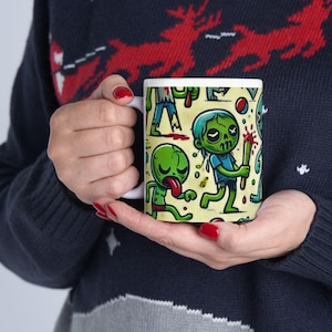 May include: A white ceramic mug with a colorful cartoon pattern of green and blue zombies. The mug has a handle and is filled with a light brown liquid.
