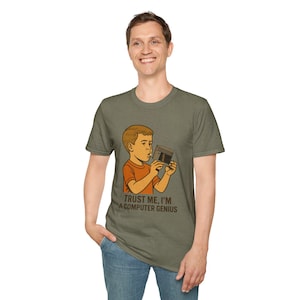 May include: A green t-shirt with a retro cartoon illustration of a boy blowing into a vintage video game cartridge. The text on the shirt reads "Trust me, I'm a computer genius."