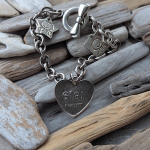 May include: A silver charm bracelet with a heart charm that says "Gigi" and other charms including a star and a bow. The bracelet is on a wooden background.