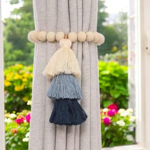 Pode incluir: Um conjunto de amarras de cortina de contas de madeira com uma borla de três camadas em creme, azul e azul escuro.