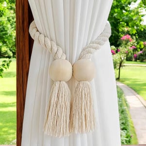 May include: Cream-coloured curtain tiebacks with a natural, rope-like design. Each tieback features a large, round wooden bead and a long, fringed tassel. The tiebacks hold white curtains, with a blurred green outdoor background.