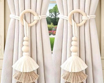 May include: Two cream-colored curtain tiebacks with wooden rings and beaded tassels. The tassels have three layers of fringe in shades of white, tan, and brown. The curtains are a light cream color, and the background shows a view of a garden.