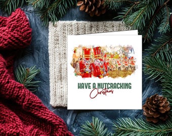 May include: A Christmas card with a watercolor illustration of nutcrackers in red uniforms. The card has the text "Have a Nutcracking Christmas" in green and red script. The card is surrounded by a red scarf, a knitted beige hat, pine needles, and a pine cone.