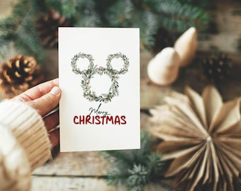May include: A Christmas card featuring a Mickey Mouse head made of green wreaths with red berries. The words "Merry Christmas" are written in red script below the design. The card is held against a rustic wooden background with pine needles and pine cones.