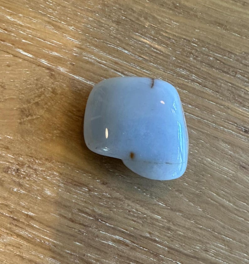 May include: A smooth, light blue gemstone with a rounded shape and a polished surface. The stone has small brown inclusions and is set against a wooden background.