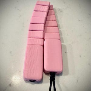 May include: Two pink, rectangular objects connected by a black cord. Each object is made of stacked, pink, rectangular blocks. The blocks have a textured surface. The cord is knotted at the base of the objects.