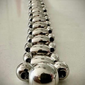 May include: A bracelet composed of a single row of polished silver-toned beads that gradually increase in size from the top to the bottom. The beads are spherical and oval in shape, featuring a highly reflective metallic surface finish throughout.