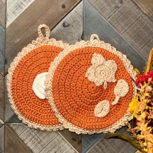May include: Two handmade crochet pot holders. The pot holders are round with an orange center and a beige scalloped edge. One has a beige flower and leaf design. Each has a loop for hanging.