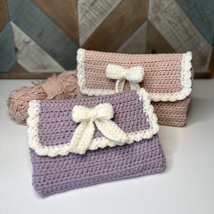 May include: Two handmade crochet clutch purses. One is lavender, the other is light pink. Both have a white bow and trim. A ball of yarn in a similar color is next to the purses. The background is a wooden chevron pattern.