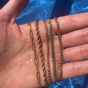 May include: Five gold-coloured chain necklaces of different styles are shown on a hand. The chains include a paperclip chain, a rope chain, a figaro chain, a herringbone chain, and a curb chain. The background is a blue water surface.