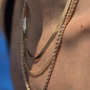 May include: A close-up of three gold necklaces layered on a person's neck. The necklaces have different designs: a flat herringbone chain, a rope chain, and a thinner chain. The skin has water droplets.