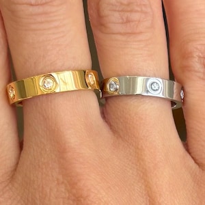 May include: Two rings, one gold and one silver, each with multiple small, clear gemstones. The gold ring is on the left, and the silver ring is on the right. Both rings are on fingers, showcasing their design and sparkle.