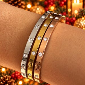 May include: Three hinged bangle bracelets in silver, gold, and rose gold tones, each featuring a smooth polished finish and a series of evenly spaced, round, clear embedded crystals along the outer surface of the metal bands.