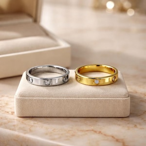 May include: Two rings, one silver and one gold, each with small, clear gemstones set into the band. The rings are displayed on a beige velvet surface, with a jewellery box in the background.