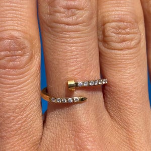May include: A gold ring with a wrap-around design. The ring has two parallel rows of small, clear gemstones. The ring is worn on a finger, showcasing its elegant and modern style.