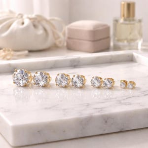 May include: A collection of gold-colored stud earrings with clear, sparkling cubic zirconia stones. The earrings are arranged in a row, decreasing in size. The earrings are displayed on a white marble surface, with a soft focus background.